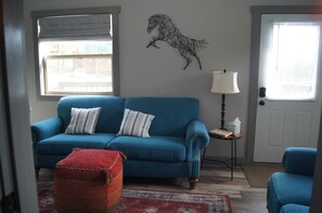 Una sala de estar con un sofá azul, un reposapiés rojo, una escultura de un caballo montado en la pared, una lámpara y una puerta con persianas.