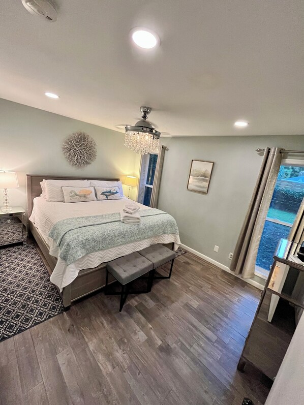 A bedroom with a bed, bedside tables, a chandelier, a bench, a view of the ocean, and a ceiling fan.