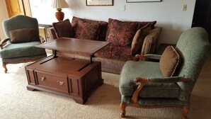 Una sala de estar con una mesa de centro marrón, dos sillones verdes, un sofá marrón con cojines estampados y una mesita de madera.