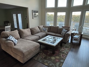 A living room with a beige sectional sofa, a glass-top coffee table, and a wooden floor. There is a window with a view of trees and a white chair in the background.