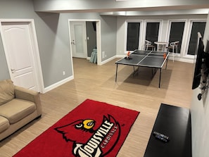 A living room with a ping pong table, a red rug with a logo, a sofa, and a TV.