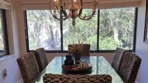 A dining area with a glass table, woven chairs, a chandelier, and a view of trees through large windows. - 3BR 2.5 BA, short walk or 5 min bike to beach w/ cart, chairs, pools, sm. dog (Hilton Head Island)