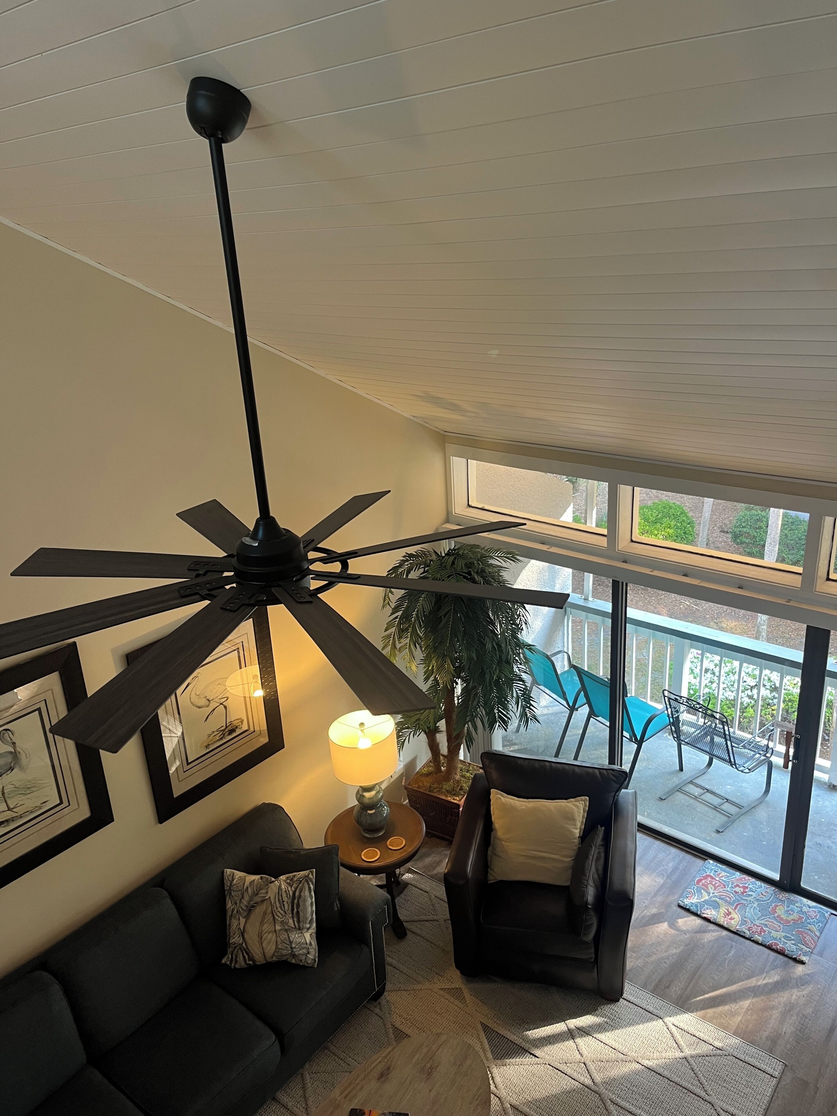 Una sala de estar moderna con ventilador de techo, un sofá negro, una mesita con lámpara y vistas a un balcón con mobiliario exterior.