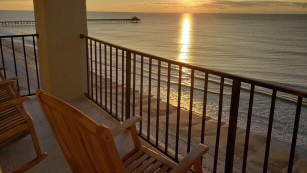 A balcony with wooden chairs and a view of the ocean at sunset.