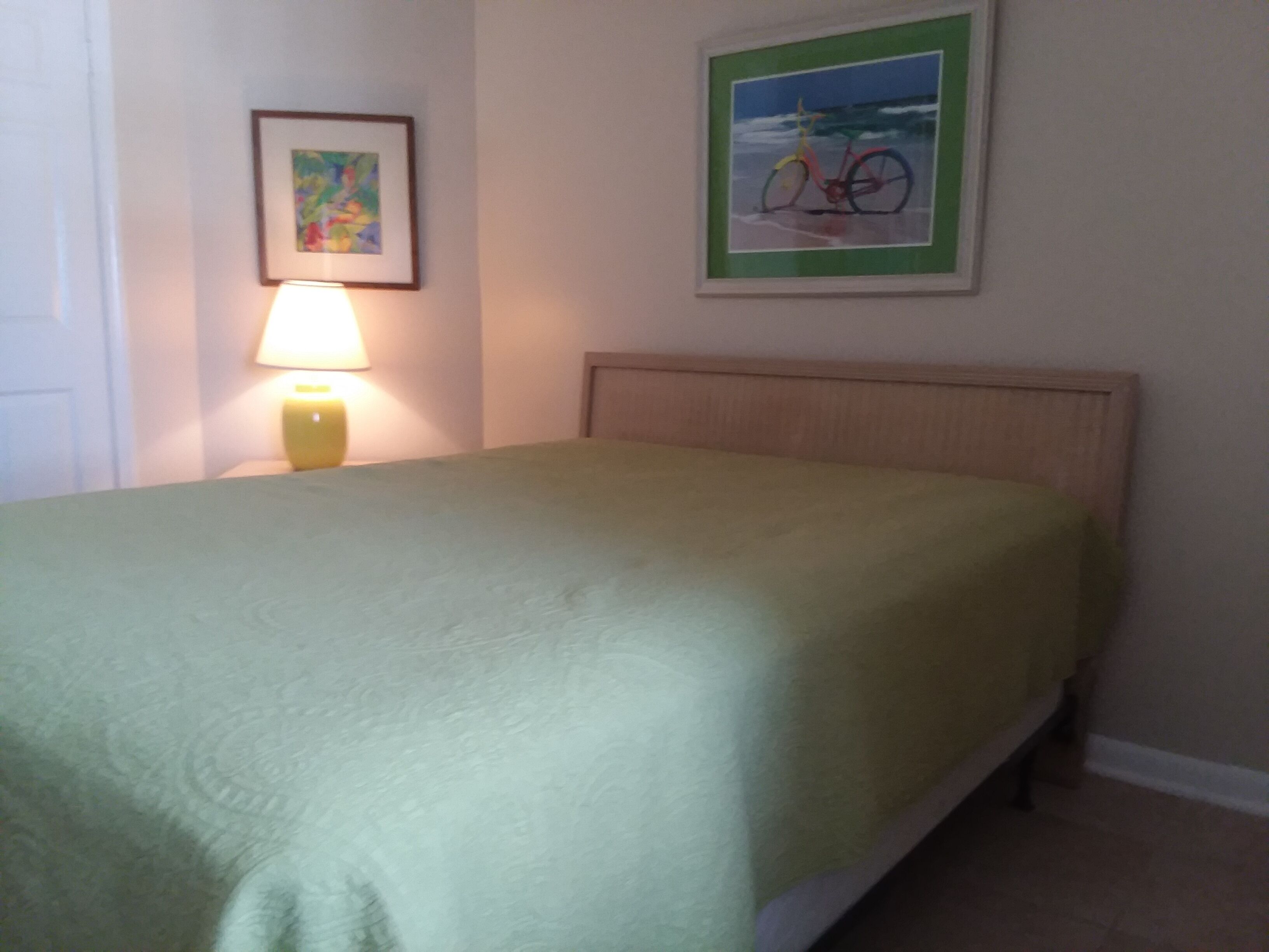 A neatly made bed with a green bedspread, a bedside lamp, and a framed picture of a bicycle on the wall.
