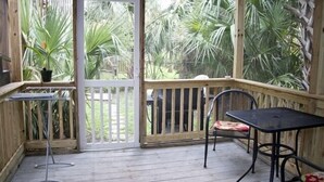 Une terrasse en bois avec une table et des chaises, une petite table ornée d’une plante, et une vue sur un jardin où poussent différentes espèces végétales.