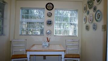 Un coin repas avec une table et des chaises blanches, deux fenêtres équipées de stores et un mur décoré d’assiettes.
