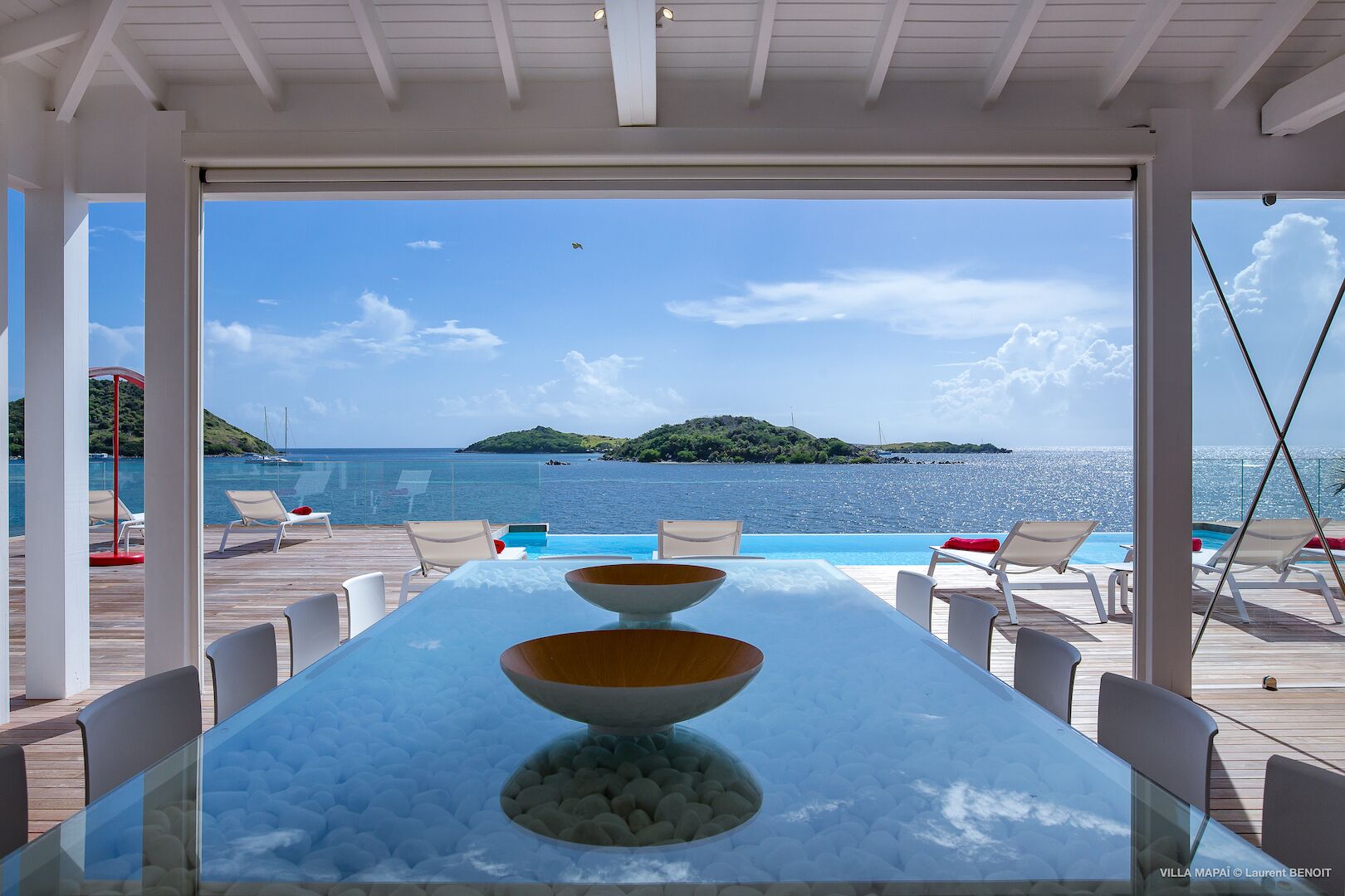 Une table avec un plateau en verre, deux bols et une chaise blanche, placée de manière à offrir une vue sur une piscine et un paysage pittoresque de la mer et d’une petite île.
