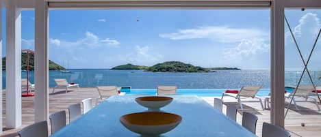 A glass-top table with two bowls and a white chair, overlooking a pool and a scenic view of the sea and a small island.