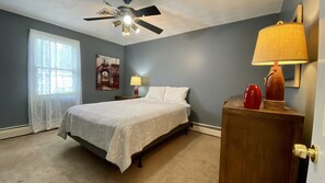A bedroom with a bed, a ceiling fan, a window with sheer curtains, a wall-mounted lamp, and a decorative vase on a wooden cabinet. - Narragansett R.I. 5 Bedrooms, 3 full bathrooms, central air on cul-de-sac (Narragansett)