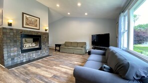 A living room with a fireplace, a sofa, a television, and a window with a view of greenery. - Narragansett R.I. 5 Bedrooms, 3 full bathrooms, central air on cul-de-sac (Narragansett)