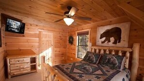 Une chambre à coucher dans un chalet en bois, avec un lit, un ventilateur de plafond, une télévision et un tableau représentant un ours accroché au mur.