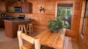 L’intérieur d’un chalet en bois comprend un coin cuisine équipé d’un four à micro-ondes, d’une cuisinière et de meubles de rangement en bois. On y trouve également une table à manger avec deux tabourets et une plante en pot.