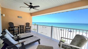 A balcony with a ceiling fan, two Adirondack chairs, a small table, and a bench. The balcony overlooks a beach with a clear view of the ocean.