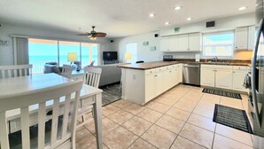 Cozinha espaçosa com armários brancos, sala de jantar com mesa e cadeiras brancas e vista para o oceano.