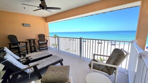 A balcony with a ceiling fan, two Adirondack chairs, a small table, and a view of the beach.