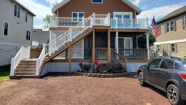 Une maison à deux étages avec un porche à l’avant, un escalier et un petit jardin agrémenté de fleurs rouges.