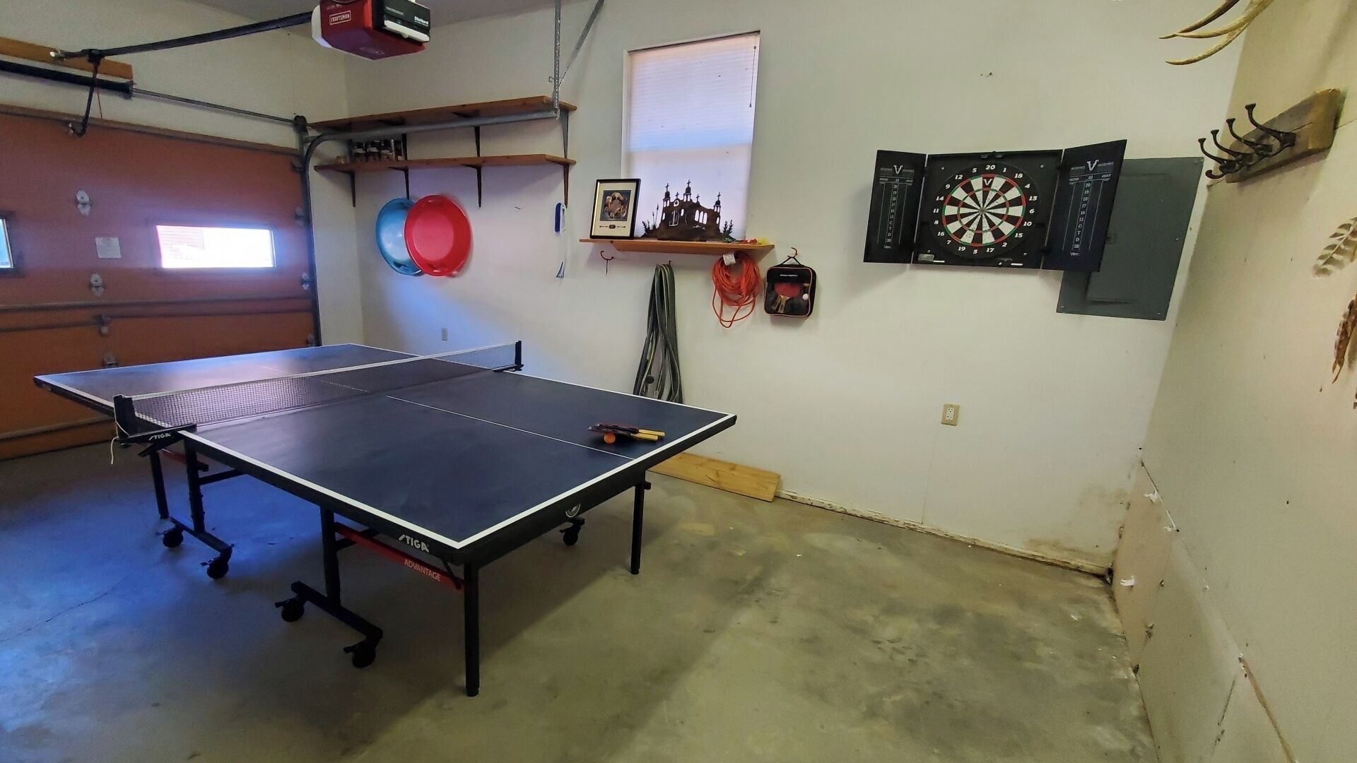 A ping pong table in a room with a dartboard, a framed picture, and a window.