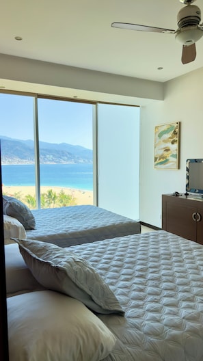 A bedroom with two beds, a ceiling fan, a desk with a computer, and a large window with a beach view.