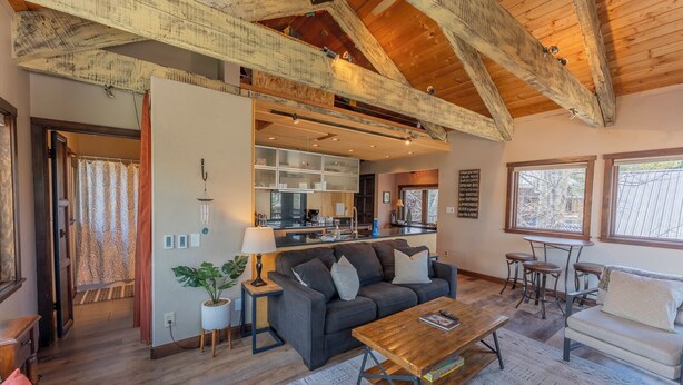 A spacious living area with a dark grey sofa, a wooden coffee table, and a small side table with a lamp. There is a kitchen area with modern appliances and a window providing natural light. - 2BR 2BA In Town, Walkable, Pet-Friendly, Fireplace, Views! (Crested Butte)