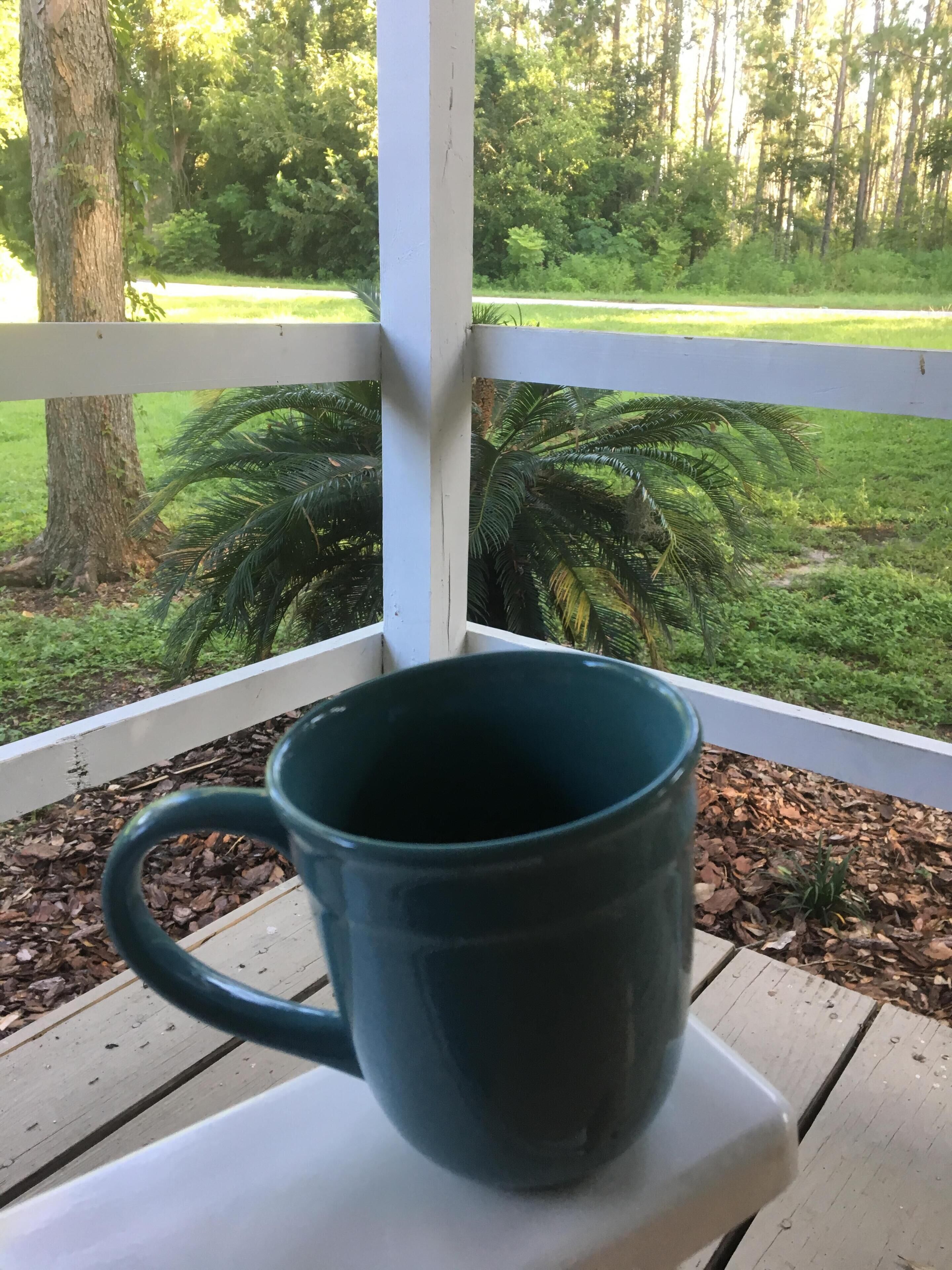 Una taza verde oscuro sobre una mesa blanca en un espacio exterior techado, con vista a un campo cubierto de césped y árboles.
