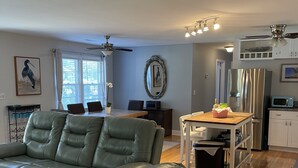 Ein Wohnzimmer mit einem grünen Ledersofa, ein Essbereich mit Holztisch und -stühlen, eine Küche mit Kühlschrank und Mikrowelle sowie ein Deckenventilator.