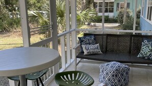 Eine überdachte Terrasse mit einem runden Tisch und zwei grünen Stühlen, einer schwarzen gewobenen Bank mit gemusterten Kissen und Blick auf ein Haus durch große Fenster.