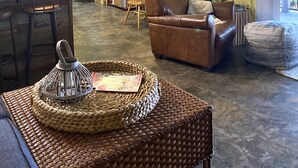 Un salon chaleureux avec un canapé en cuir marron, une table à manger avec des chaises, un coin cuisine doté d’un ventilateur de plafond et une grande fenêtre avec des stores.