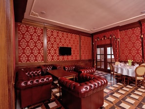 Una habitación con piso a cuadros, una mesa grande, asientos tapizados en rojo y un televisor montado en la pared.