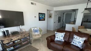 Un soggiorno con un divano in pelle marrone, un tavolino da caffè in vetro con un pesce, una TV su un supporto in legno e una cucina sullo sfondo.