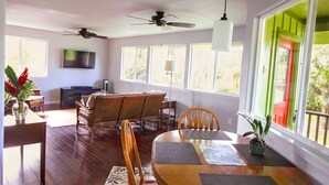 A spacious dining area with a wooden table and chairs, a ceiling fan, and large windows with a view of greenery. - Secluded garden bungalow in the jungle with air conditioning. (Keaau)