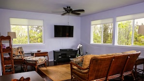 A living room with a flat-screen TV, wooden furniture, and large windows with blinds. - Secluded garden bungalow in the jungle with air conditioning. (Keaau)