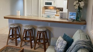 A modern kitchen with white cabinets, a microwave, and a stove. There are three wooden stools and a glass-top coffee table. A grey sofa with patterned cushions is in the living area. - BLUE / GREEN PARADISE WITH A PRIVATE BEACH!!! (Tavernier)