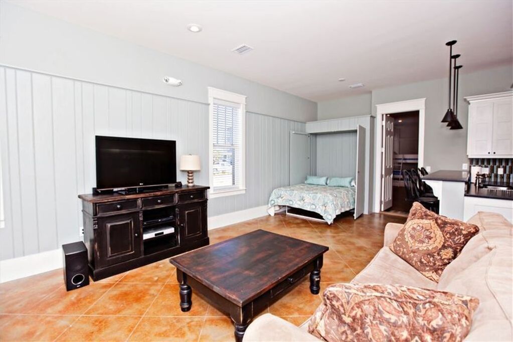 A living room with a TV, sofa, coffee table, and a kitchen area in the background.