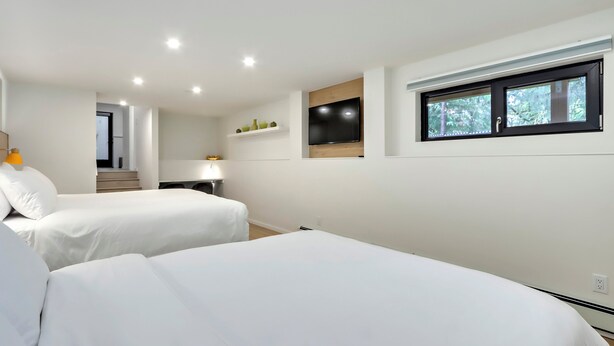 Une chambre d’hôtel moderne avec deux lits, un téléviseur fixé au mur, une fenêtre donnant sur des arbres et une petite étagère avec des objets décoratifs.