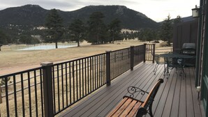 Una terraza de madera con un banco y una mesa, con vista a un área cubierta de césped con árboles y una masa de agua, con montañas al fondo.