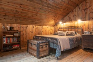 A cozy cabin-style bedroom with a wooden ceiling, a metal bed frame, a bedside table with a radio, and a bookshelf filled with books.
