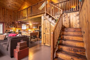 A rustic cabin interior with a wooden staircase, a cozy living area with a sofa and a dining table, and a kitchen with wooden cabinets.