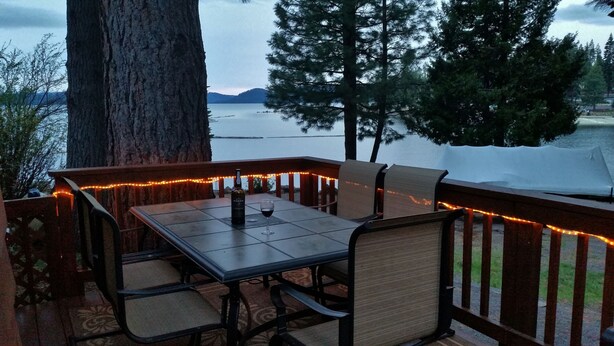 Una terraza exterior con mesa y sillas, luces colgantes y vistas a un lago y montañas.
