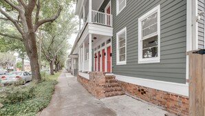A residential street with a two-story house featuring a brick foundation, a porch with white railings, and red doors. - Gwinnett St Row: Historic 3 BR 2BA - Balcony - Free St Parking - Near Forsyth Pk (Savannah)