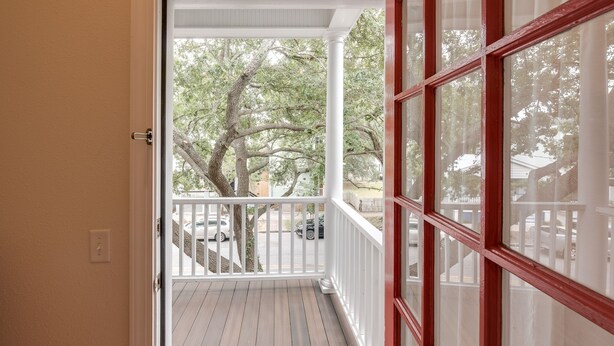 A covered porch with a wooden floor and white railings, leading to a view of a residential street with cars and trees. - Gwinnett St Row: Historic 3 BR 2BA - Balcony - Free St Parking - Near Forsyth Pk (Savannah)