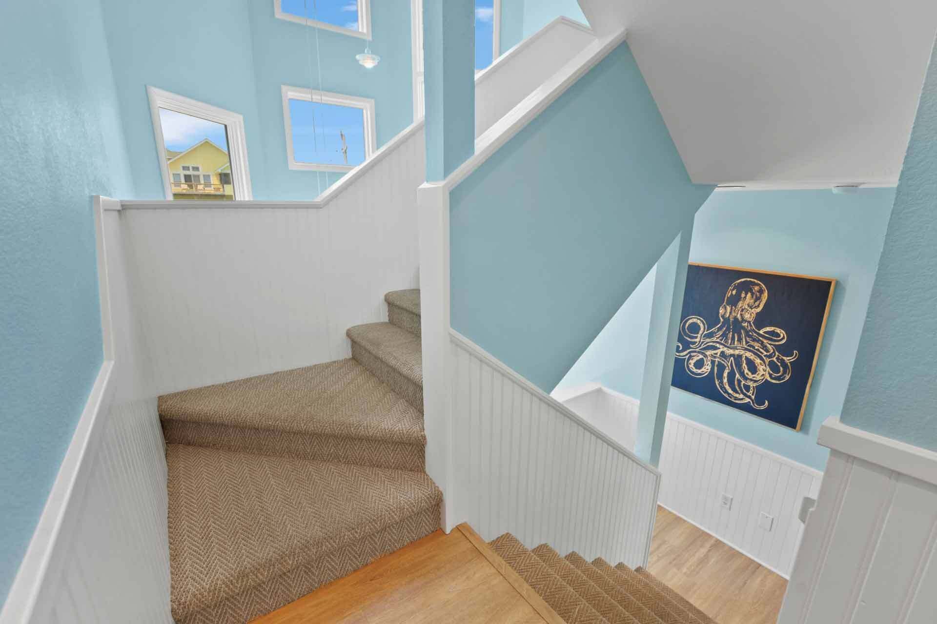 A staircase with a herringbone carpet and white railings, leading to a room with light blue walls and a framed artwork of an octopus.