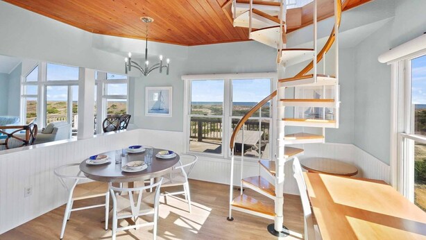 A spacious room with a wooden ceiling, a spiral staircase, and large windows offering a view of the beach. - Oceanfront Dog Friendly Home with Pool, Hot Tub, EV Charger, and Home Theater (Salvo)
