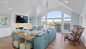 A spacious living room with a large sectional sofa, a wooden coffee table, and a TV mounted on the wall. There are two chairs and a wicker chair near the windows. - Oceanfront Dog Friendly Home with Pool, Hot Tub, EV Charger, and Home Theater (Salvo)