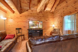 A cozy log cabin interior with a leather sofa, a wooden chair, a fireplace, and a television mounted on the wall. - Blue Rose Cabins - Oleander Cabin (Logan)