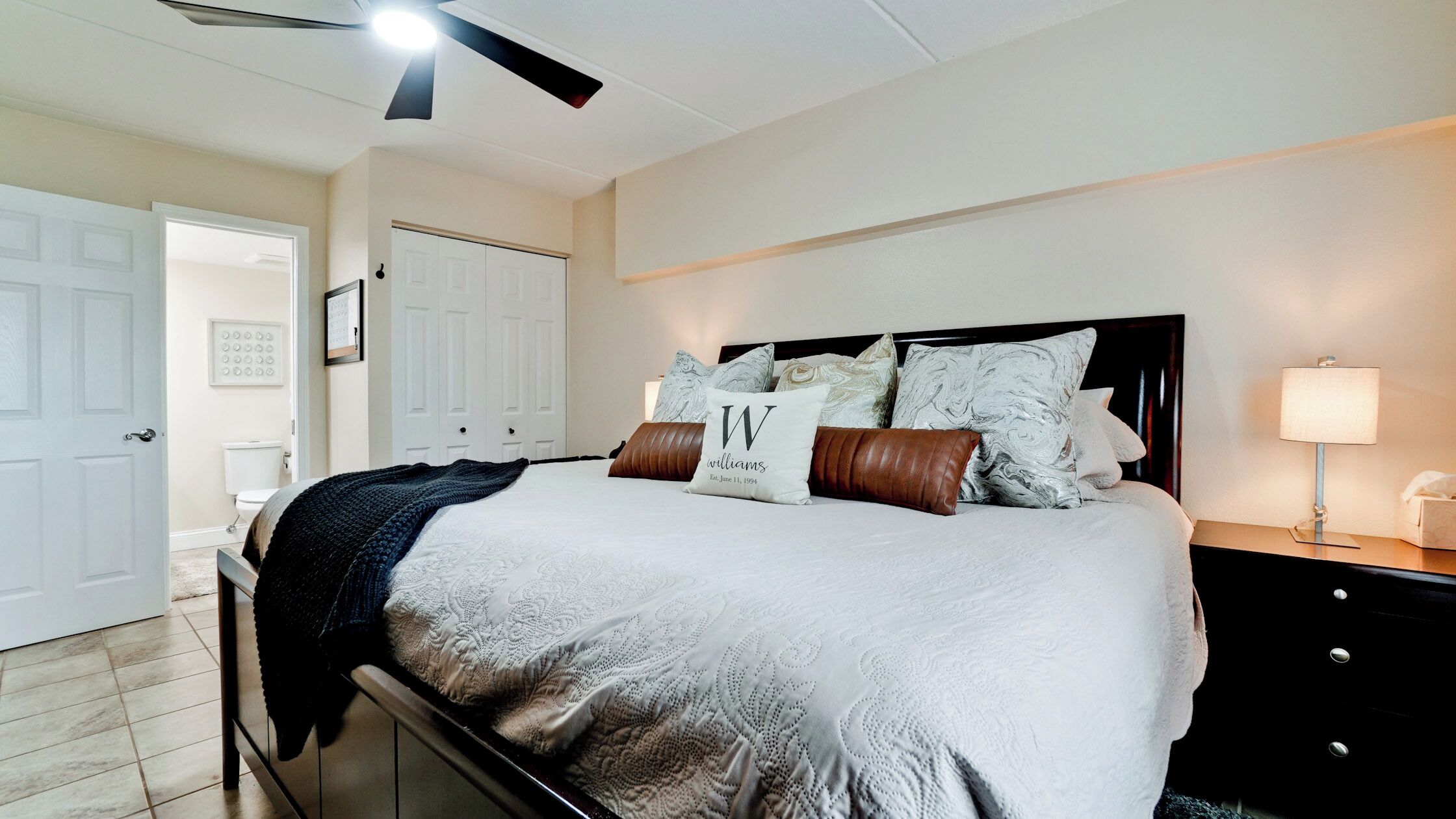 Een netjes ingerichte slaapkamer met een bed, nachtkastje met lamp, garderobe en plafondventilator.