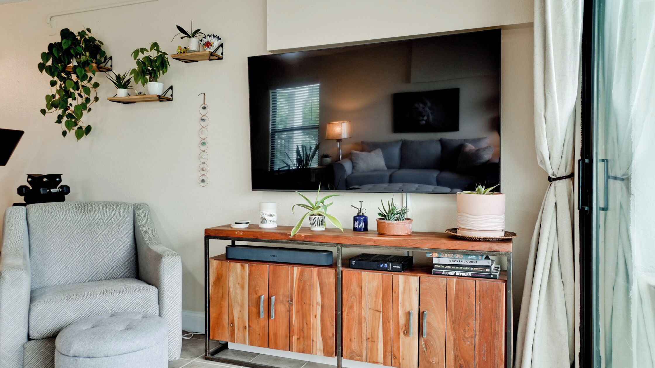 A living room with a grey armchair, a wooden cabinet with books, a TV, and wall-mounted shelves with plants.