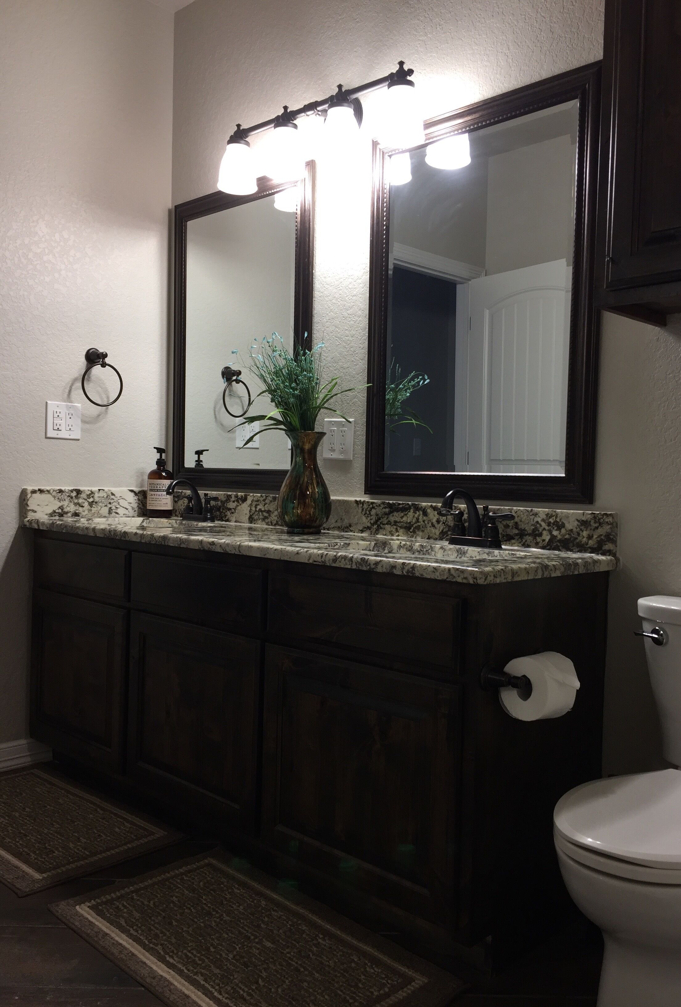 A bathroom with a dark wood vanity, a large mirror with lights, a vase with green plants, and a toilet.