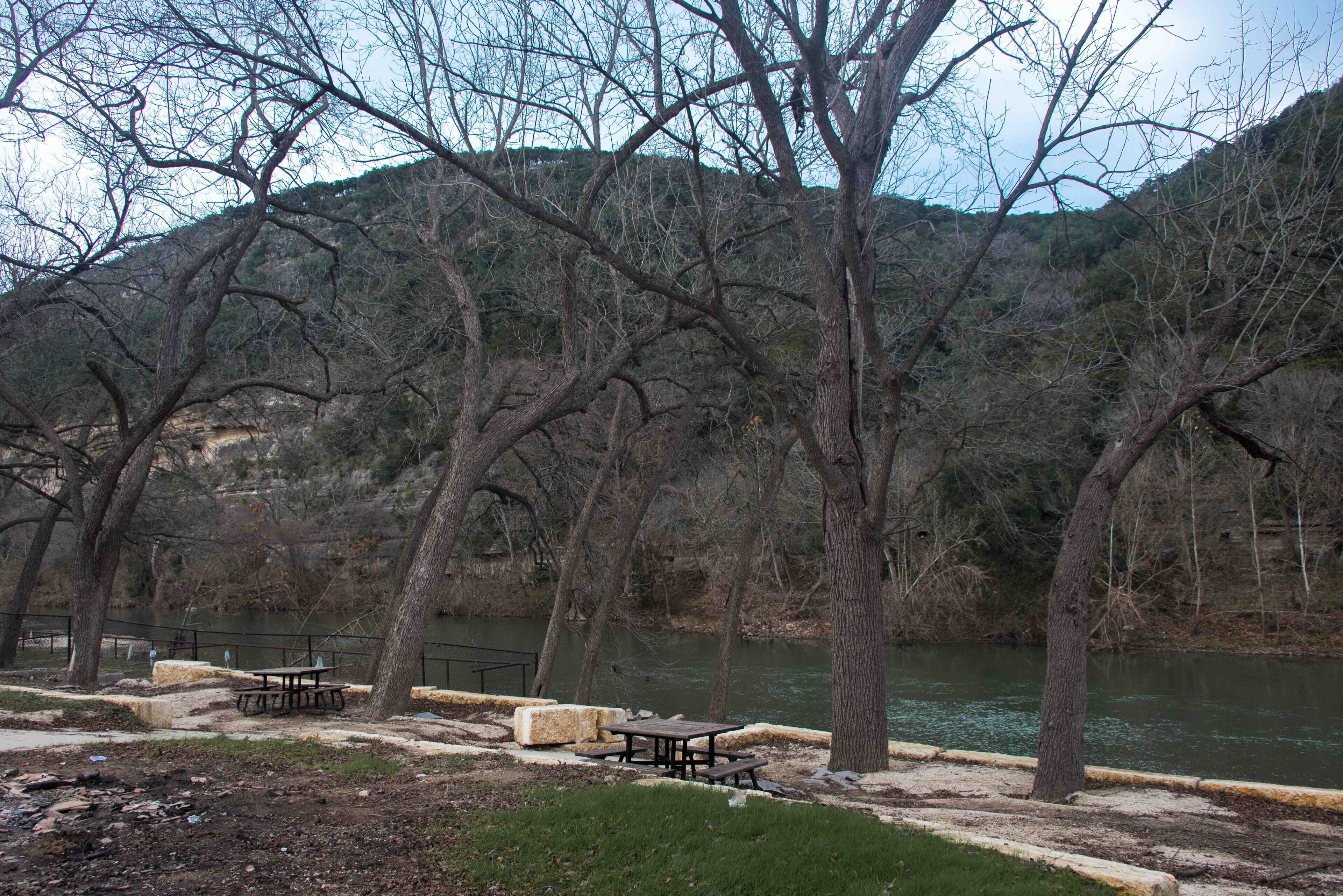A riverside park with picnic tables and benches, surrounded by trees and hills.