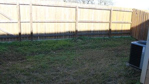 A backyard with a wooden fence, a grassy area, and an air conditioning unit.